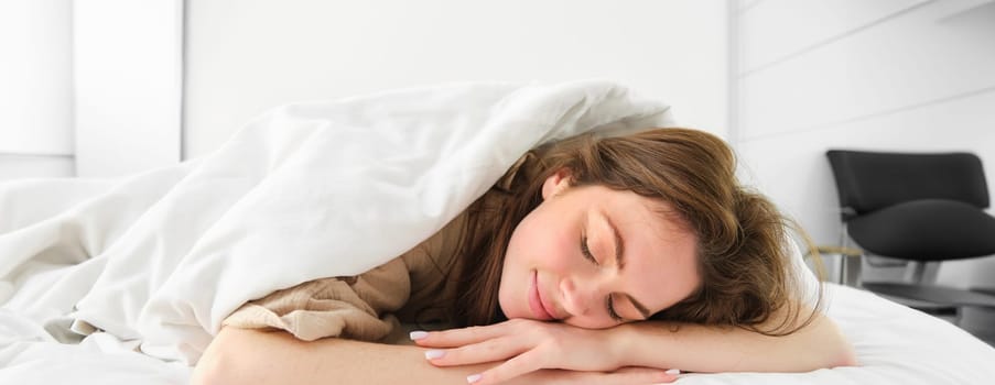 Cute young woman lying under blanket and smiling, sleeping, having a good morning nap, lazy day in bed. Lifestyle and home concept