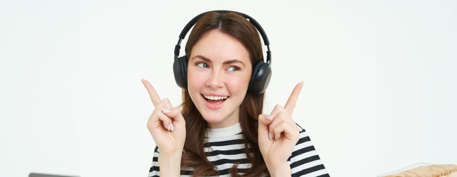 Dancing girl in wireless headphones pointing at empty space for music store promo text, showing advertisement, isolated over white background.