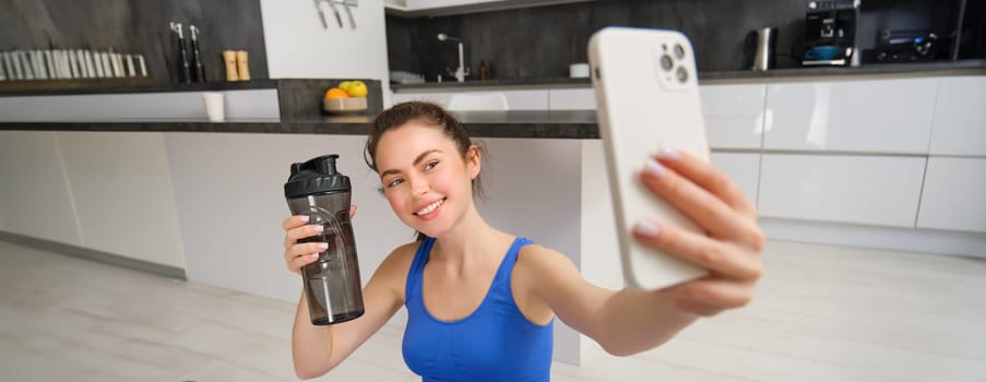 Portrait of sportswoman takes selfie with water bottle in living room, holds smartphone and poses for photo while doing fitness workout.