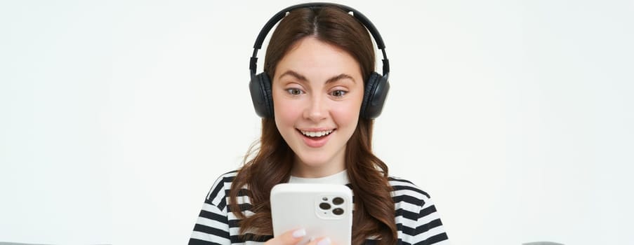 Young woman with surprised face, looking at smartphone screen, reacts amazed, looks with happiness at mobile phone, standing in wireless headphones, white background.