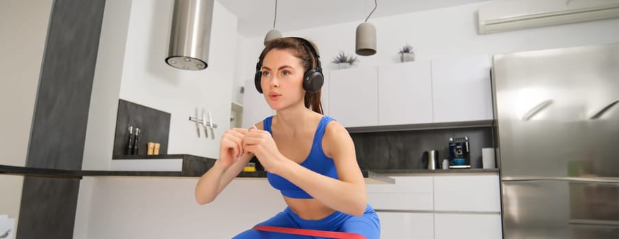 Image of beautiful fitness girl in headphones, does squats exercises with elastic band, resistance training, standing on yoga mat.