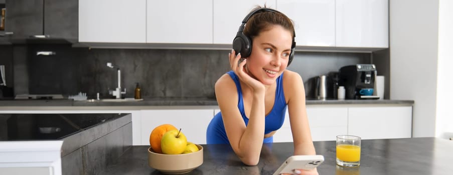 Portrait of beautiful young woman in fitness sportswear, listening music in headphones, holding smartphone, drinking juice in kitchen after workout training session.