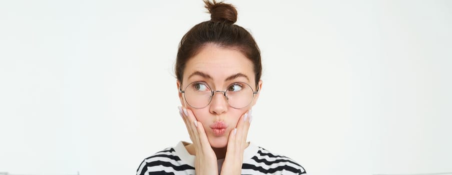 Close up portrait of cute girl in glasses, holds hands on face and puckers lips, looks aside, makes silly face expression, stands over white background.
