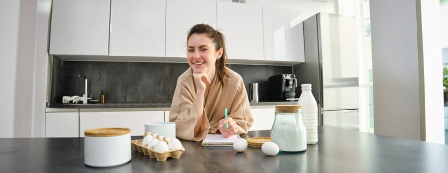 Attractive young cheerful girl baking at the kitchen, making dough, holding recipe book, having ideas.