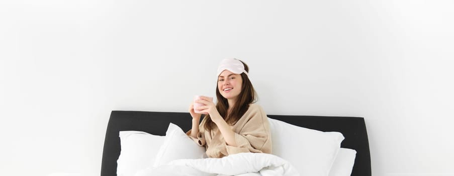 Portrait of beautiful woman enjoys her morning cup of coffee from bed, sitting in bedroom wearing sleeping mask and pyjamas, relaxing.