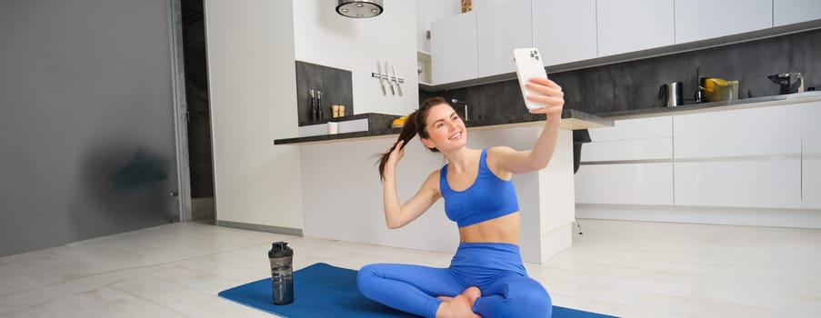 Sport and lifestyle concept. Young woman doing workout at home on yoga mat, makes fitness vlog with smartphone, takes selfie on mobile phone.