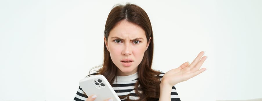 Portrait of confused young woman, looks frustrated, shrugs shoulders, hold mobile phone in one hand, white isolated background.
