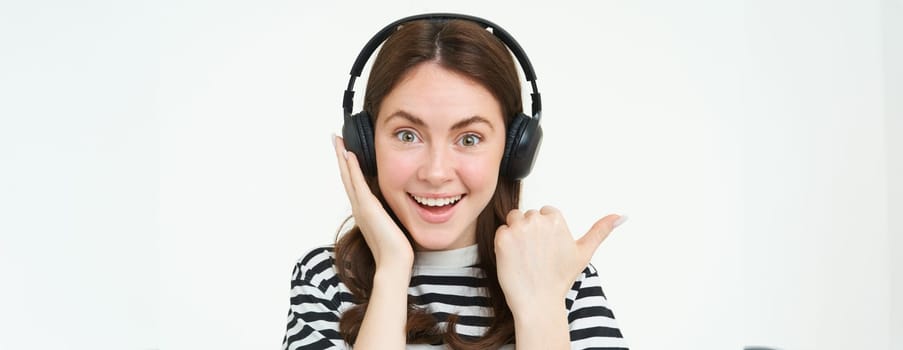 Portrait of excited woman in wireless headphones, showing awesome news, pointing left, standing over white background.