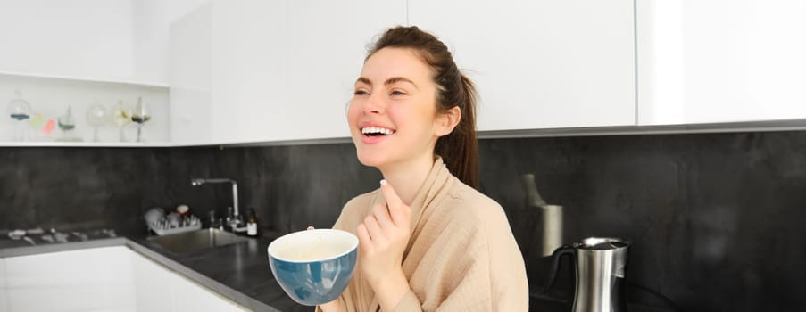 Portrait of good-looking young woman starting her day with cup of coffee, standing in the kitchen and drinking cappuccino from big mug, enjoying favourite drink in the morning.