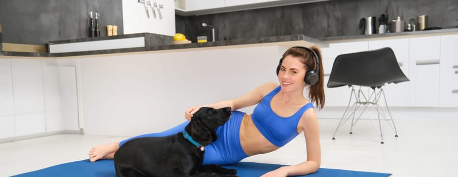 Portrait of beautiful young fitness woman, laying on yoga mat , workout at home, playing with her dog while doing sports. Lifestyle and wellbeing concept