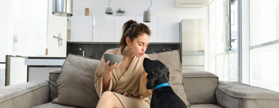 Happy young woman in bathrobe, enjoys spending time with her dog at home, eating cereals, having breakfast with puppy, sitting on sofa.