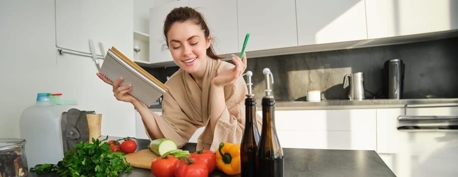 Portrait of woman checking recipe notes in notebook, standing in kitchen with vegetables, cooking food, preparing delicious salad from tomatoes, parsley and olive oil.