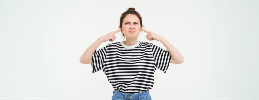 Image of annoyed, bothered woman plugs, shuts her ears with fingers, disturbed by loud noise, stands over white background.