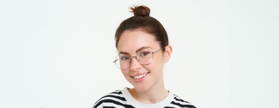 Close up portrait of cute young woman in glasses, looking at camera and smiling, trying on new spectacles, standing over white background.