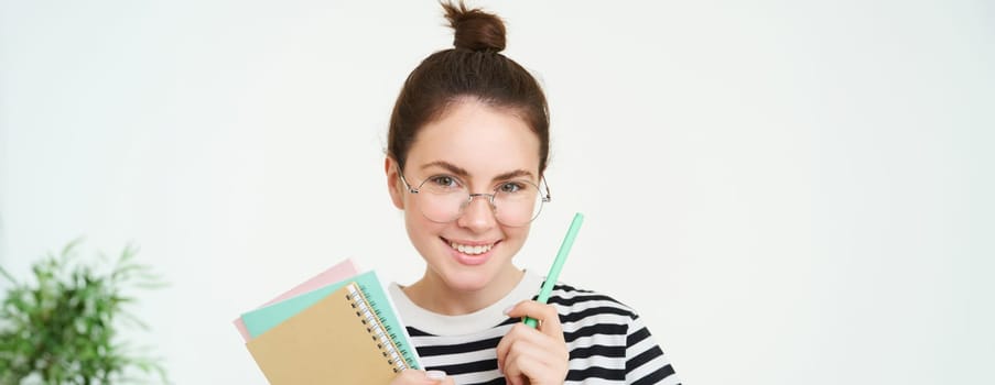 Close up portrait of young woman, tutor, online teacher in glasses, holding pen and documents, carries notebooks, stands over white background.