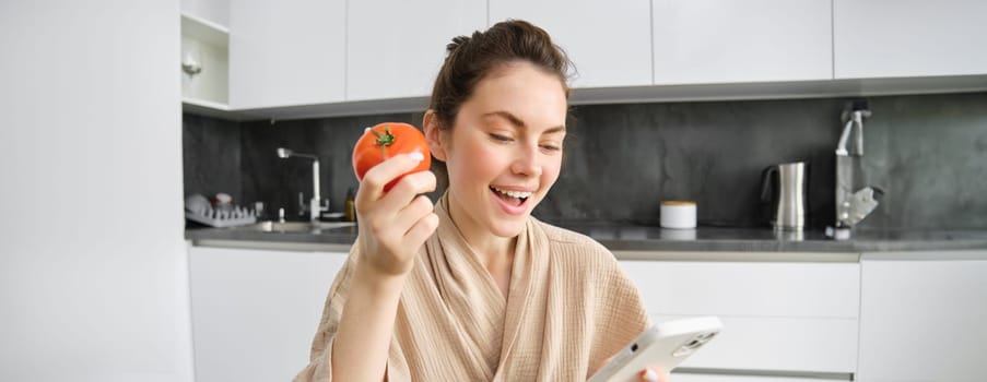 Close up portrait of beautiful young woman in the kitchen, holding tomato and smartphone, buys fresh vegetables online, orders groceries delivery on mobile phone application.