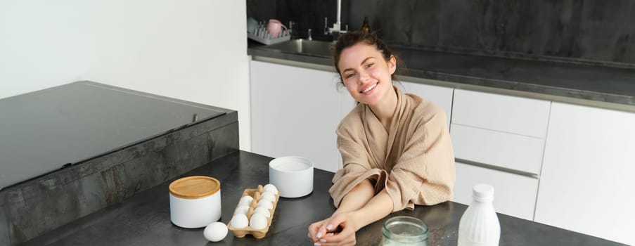 Attractive young cheerful girl baking at the kitchen, making dough, holding recipe book, having ideas.