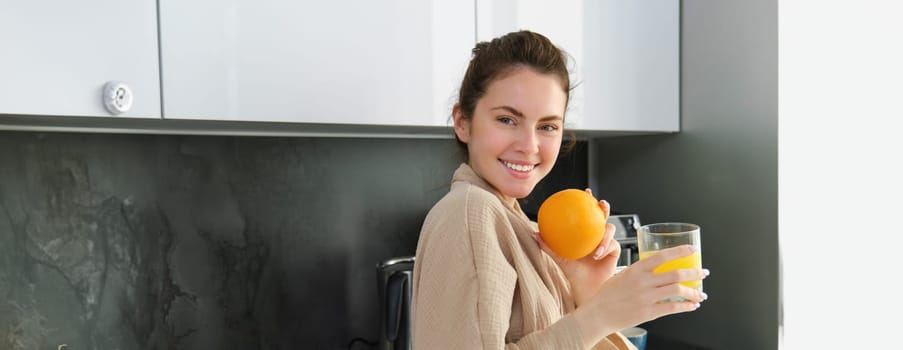 Portrait of happy woman in kitchen, wearing bathrobe, drinking orange juice, freshly squeezed drink, smiling and laughing, food and drink concept.