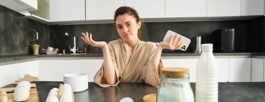 Attractive young cheerful girl baking at the kitchen, making dough, holding recipe book, having ideas.