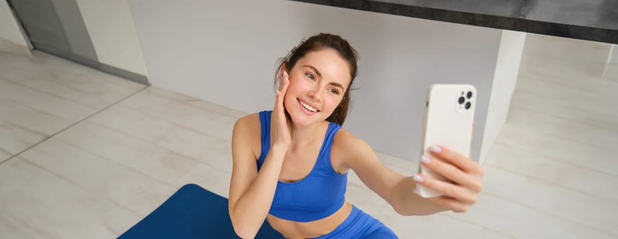 Image of beautiful, atheltic girl sits on yoga mat at home with smartphone, takes selfie and workout, does fitness exercises.