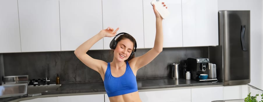 Portrait of happy, carefree fitness woman, wearing clothes for sport, dancing in kitchen with smartphone, wearing wireless headphones, listening music.