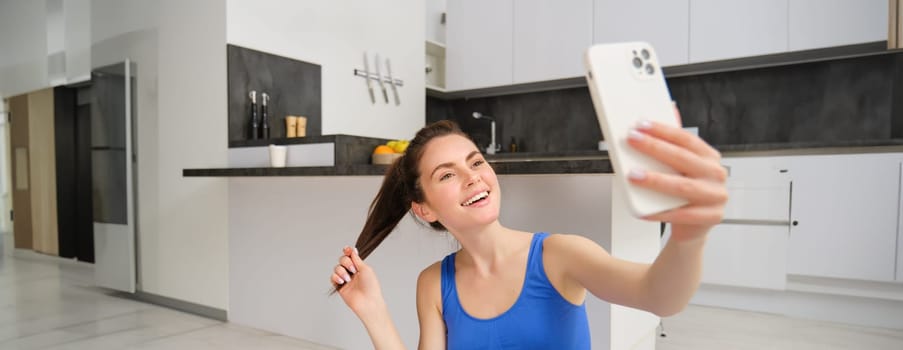 Close up portrait of young sporty woman, fitness girl takes selfie at home while doing her workout, posing on yoga mat, holds smartphone and smiles.