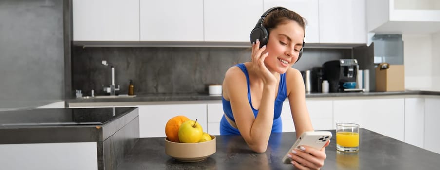 Image of happy, stylish young sports woman, standing in kitchen and drinking orange juice, listening music in headphones, using smartphone app.