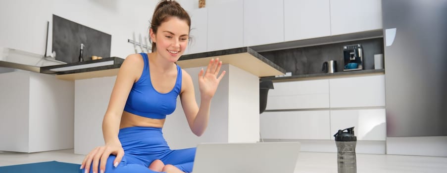 Portrait of fitness instructor, woman connects to online training session, waves hand at laptop, teaching yoga workout from home, sitting on rubber mat.
