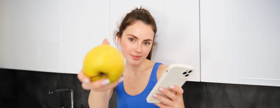 Image of beautiful, sportswoman sitting in kitchen with smartphone, offering you an apple, eating fruits, wearing sportsbra and leggings.