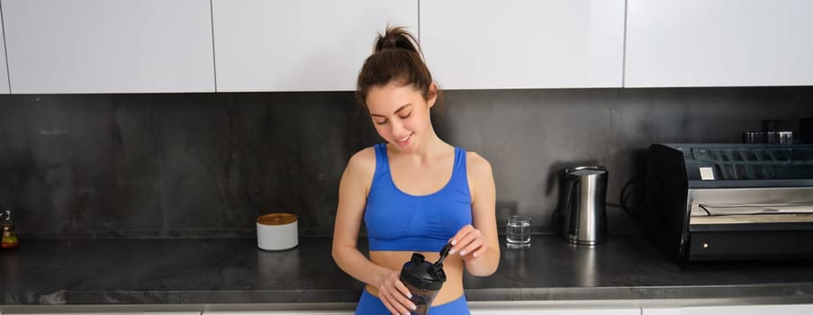 Image of young woman preparing dietary supplement, protein in her shaker bottle, wearing fitness clothing, standing in kitchen.