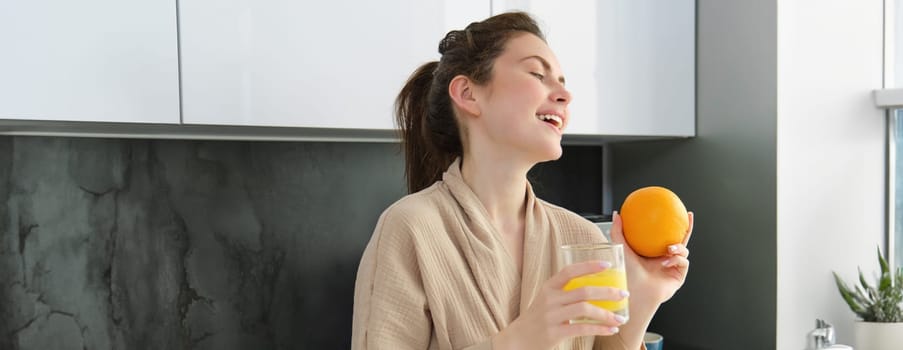 Portrait of happy woman in kitchen, wearing bathrobe, drinking orange juice, freshly squeezed drink, smiling and laughing, food and drink concept.