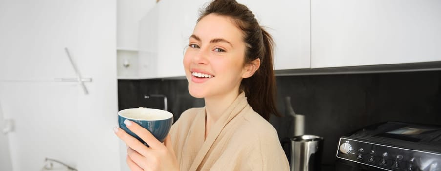 Portrait of good-looking young woman starting her day with cup of coffee, standing in the kitchen and drinking cappuccino from big mug, enjoying favourite drink in the morning.