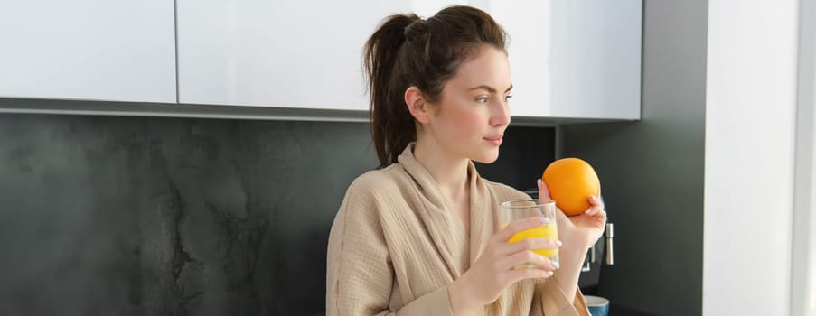 Beautiful young woman in bathrobe, drinking homemade orange juice, smiling and laughing, standing near worktop in kitchen.