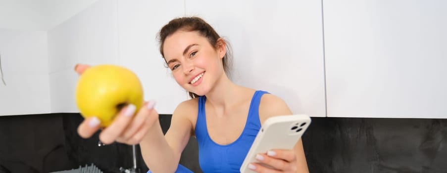 Portrait of beautiful, smiling young fitness woman, offering you an apple, eating healthy snack, holding a fruit, sitting in kitchen.