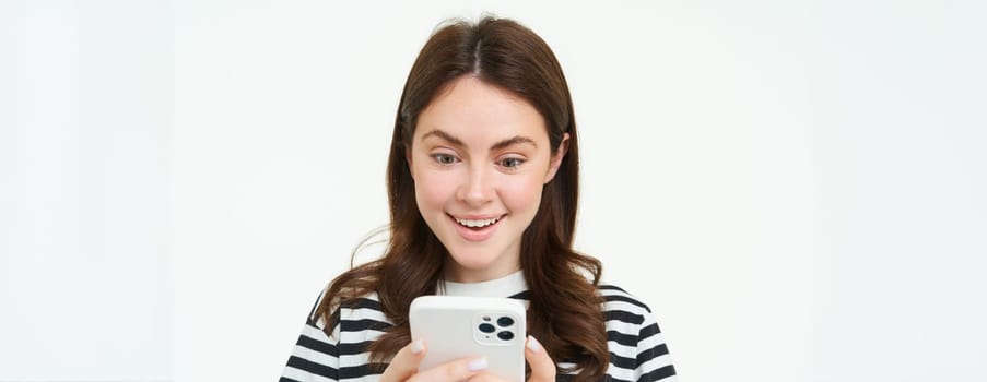 Portrait of woman looking surprised at mobile phone screen, reads amazing promo in online store, standing over white background.