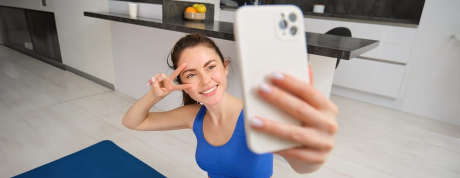 Portrait of girl takes selfie on smartphone while does her workout training. Young woman does her exercises at home, takes selfie for sports blog, sits in living room.