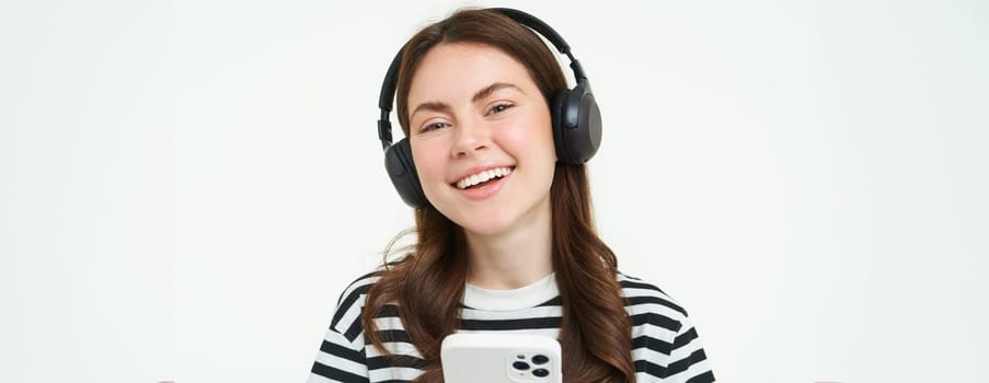 Portrait of beautiful young woman listens to music in wireless headphones, uses her mobile phone, smiles and looks happy, isolated on white background.