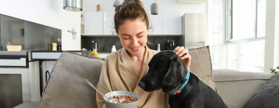 Happy young woman in bathrobe, enjoys spending time with her dog at home, eating cereals, having breakfast with puppy, sitting on sofa.