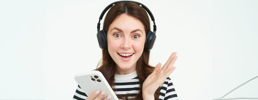 Cheerful, beautiful young woman using smartphone app, celebrating, looking happy while playing with mobile phone, listening music in wireless headphones.