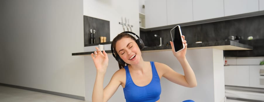 Cheerful brunette girl does fitness at home, wears wireless headphones and dances, holds smartphone, does workout indoors on floor, wears blue activewear.