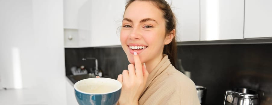 Close up portrait of attractive girl drinking coffee, holding cup with morning cappuccino and smiling, having a mug of delicious drink in the kitchen, wearing comfortable bathrobe.