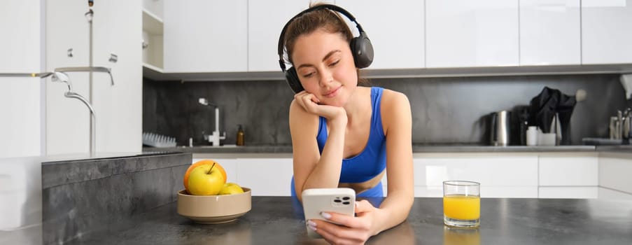 Lifestyle and workout. Young smiling woman in headphones, standing in kitchen with smartphone, drinking orange juice and listening music, heading over to gym, going jogging.