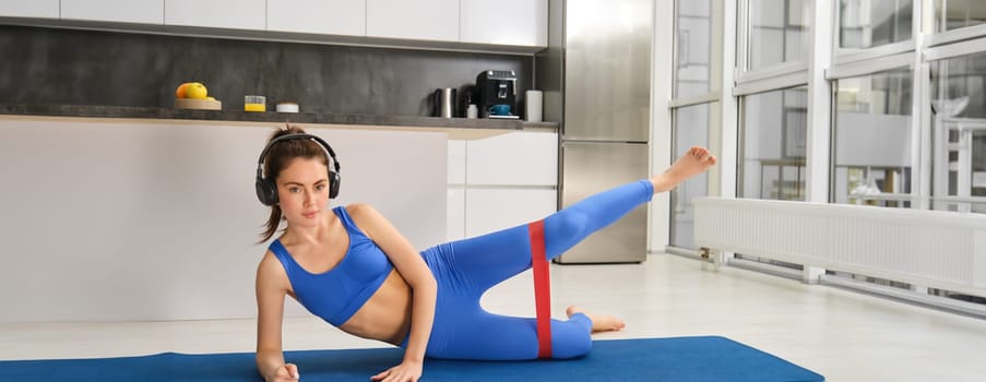 Image of sporty girl doing aerobics, using resistance band on legs, workout at home, listens to music in headphones.