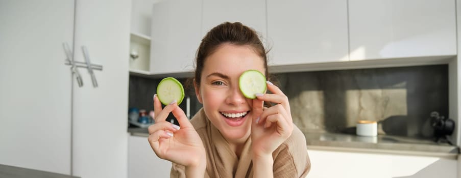 Funny, happy girl eating healthy, holding zucchini, chopping vegetables for healthy meal in the kitchen, posing in bathrobe.