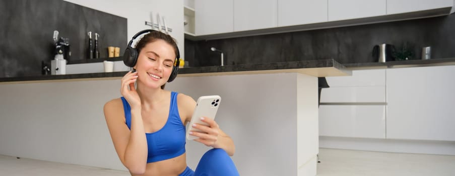 Portrait of young happy woman does workout in headphones, listens to music while doing yoga, sitting on rubber mat in blue sportswear.