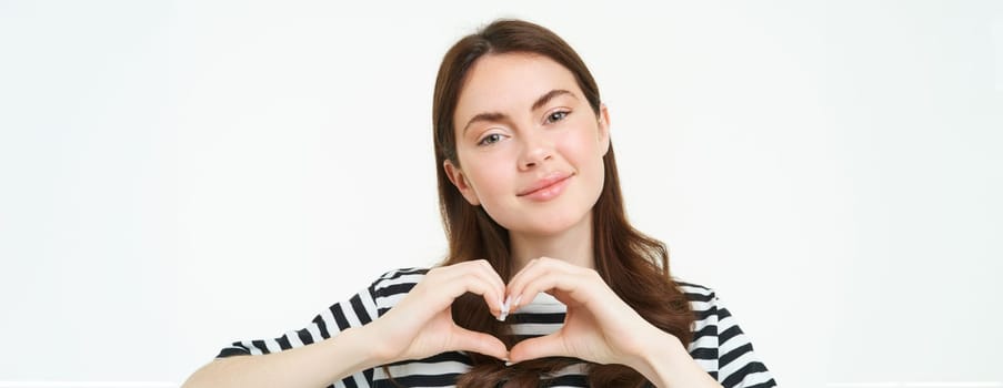 Close up portrait of cute young woman shows heart sign, gazing at camera with love and care, standing over white background.