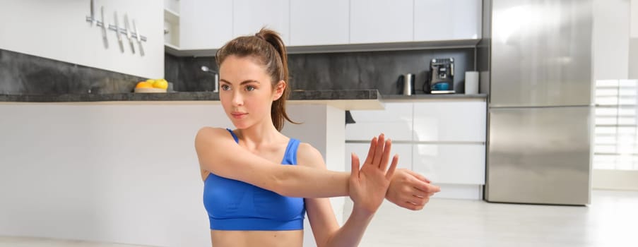 Image of fitness girl concentrates on workout, stretches hands before training session at home, follows online gym instructions.