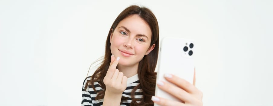 Portrait of beautiful woman showing heart sign and posing for selfie, taking picture of herself on smartphone app, posing near something cute, white background.
