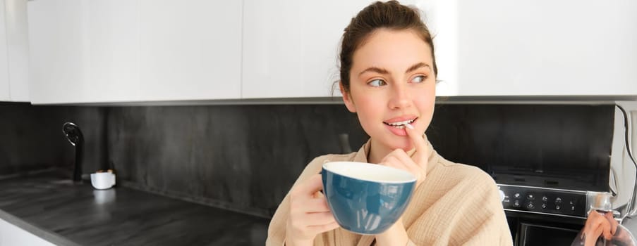 Close up portrait of attractive girl drinking coffee, holding cup with morning cappuccino and smiling, having a mug of delicious drink in the kitchen, wearing comfortable bathrobe.