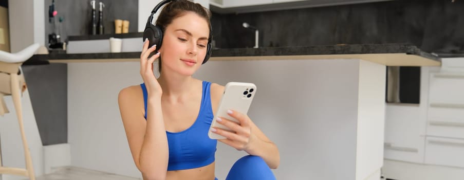 Portrait of woman in headphones, looking at smartphone, sitting on rubber mat and choosing music, doing yoga, workout training.